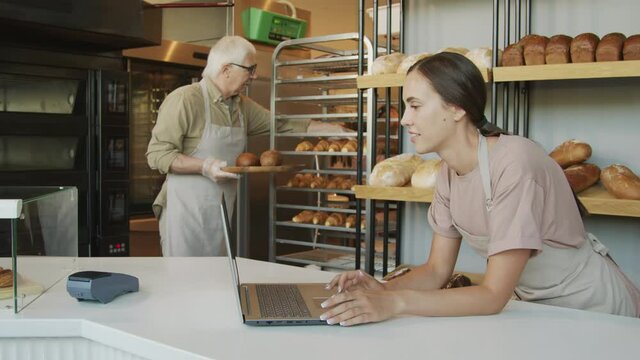 Handheld slowmo shot of senior baker or bakers assistant in apron putting fresh bread on tray and placing it on counter while looking at female counter attendant with laptop