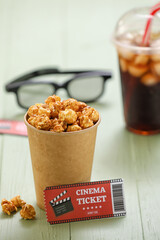 Cup with caramel popcorn and cinema ticket on color wooden background, closeup