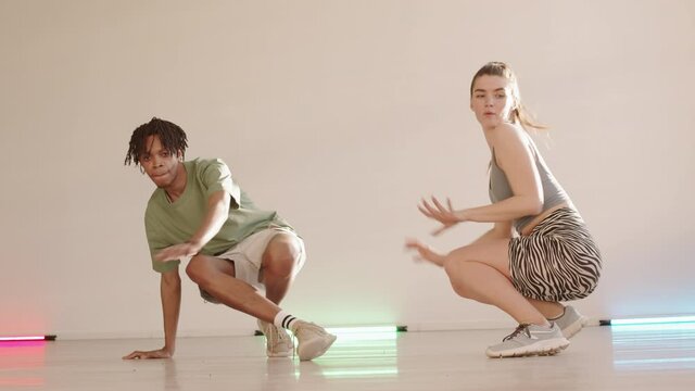 Full POV of cheerful Caucasian woman dancing in squat position, young African man dancing breakdance in studio