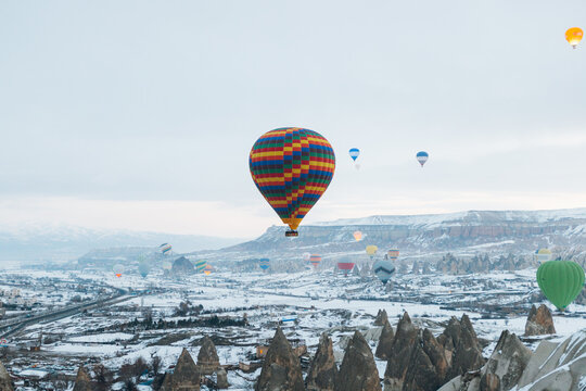 Colorful Air Balloons Flying In Gray Sky Over Countryside Against Misty Mountains At Horizon In Winter