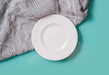 The white plate stands on a turquoise glossy table and a gray woolen knitted fabric, a top view.