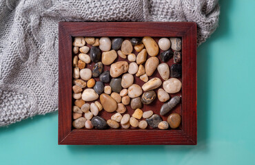 A wooden frame with sea pebbles lies on a turquoise background and a knitted plaid, a view from above. - obrazy, fototapety, plakaty