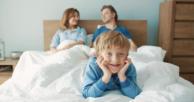 Portrait of little happy cute blond boy lying in morning on bed with his parents and smiling looking at camera.