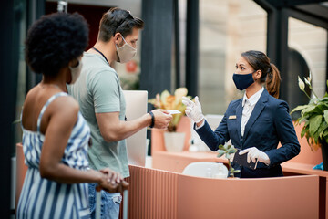 Couple with face masks communicating with receptionist while paying for their hotel stay with...