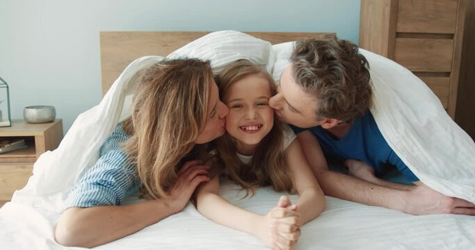 Portrait of happy caucasian couple lying in bed with cute daughter and kissing her on cheeks. Concept of parental love.