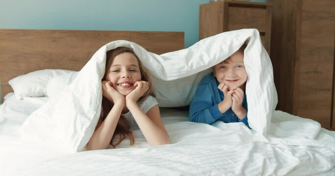 Portrait of happy little brother and sister having fun in the morning in bed hiding and appearing under blanket.
