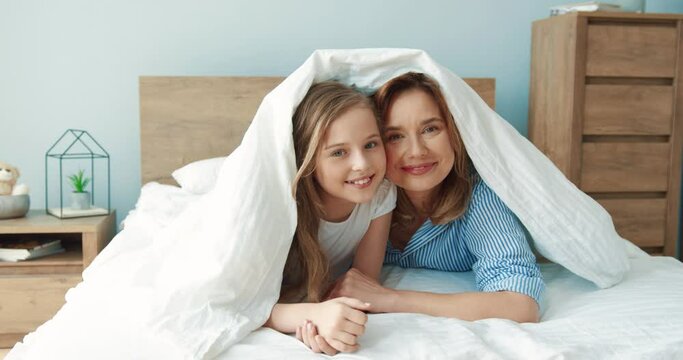 Portrait of happy mother and daughter lying on bed in the morning hiding under blanket looking at camera smiling.