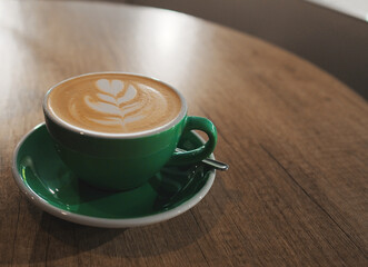 a green cappuccino cup with a saucer and a spoon on a round wooden table in a cafe or restaurant. large window on the background. milk foam with a pattern on coffee