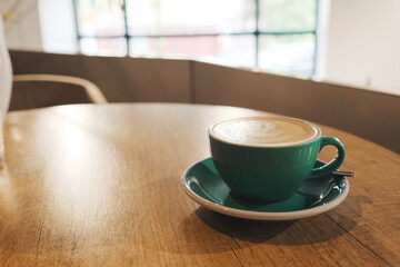 a green cappuccino cup with a saucer and a spoon on a round wooden table in a cafe or restaurant. large window on the background. milk foam with a pattern on coffee