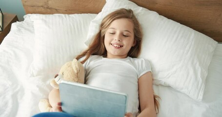 Little cute girl lying in bed with teddy bear and is surprised to see surprise on tablet.