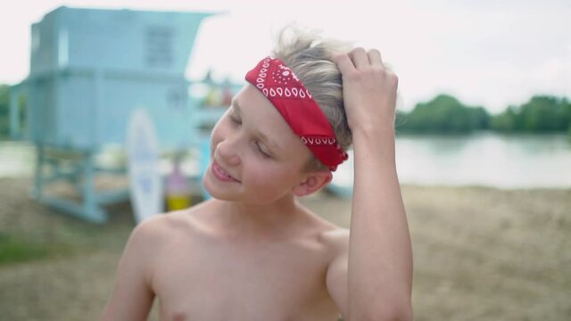 Cute blond teen boy with red headband touching his hair and posing at the sand beach against blue lifeguard tower.