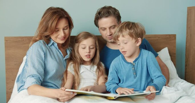Happy children and parents lying at home in bed together looking at interesting book with beautiful pictures, solving quests and secrets.