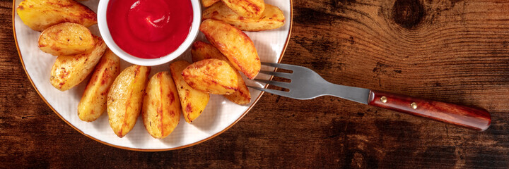 Baked potato wedges and ketchup panorama with a fork, shot from above on a dark rustic wooden background