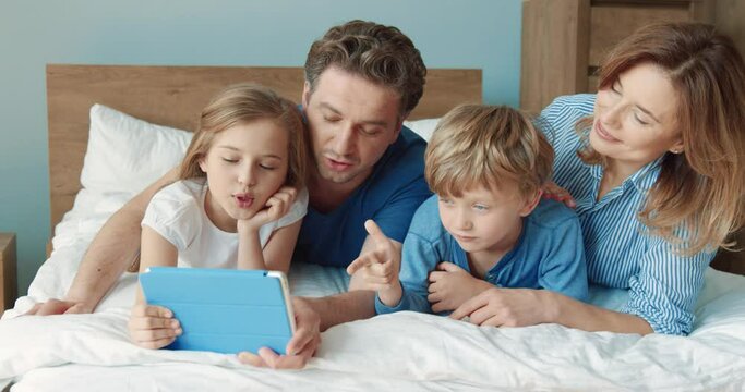 Happy parents with two children spend free morning time together lying at home on bed watching movie on tablet.