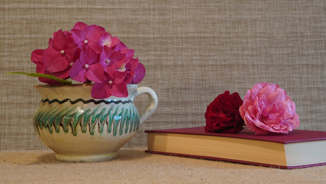 Closed book with red covers and pink and red roses and hydrangea on beige background with copy space. Traditional romanian hand painted clay vase.
