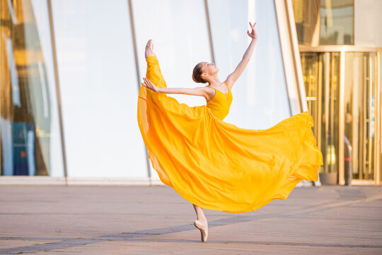 Fototapeta young ballerina in a long flying yellow dress is dancing against the backdrop of cityscape