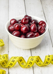 Ripe fresh cherries in a white dish, measuring tape on the background of a light brick wall. Concept of healthy food, diet. Side view. Vertical photo.