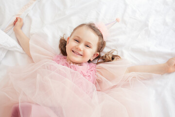Happy child girl in pink festive dress and birthday cap lies on bed spread arms looking at camera and smiling. photo above