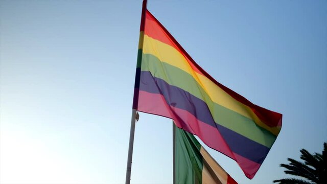 Slow motion pan to right of a beautiful pride flag waving in the wind, other flag waving in background with clear blue sky.