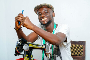 image of cheerful african man with both hands on sewing machine, holding smart phone, measuring...