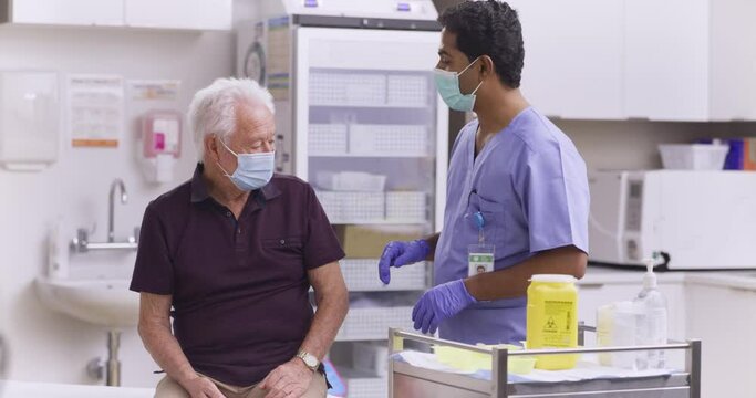 A Wide Shot Of An Indian Male Medical Nurse Administering A Covid-19, SARS-CoV-2 Vaccine Injection With A Needle and Syringe, To An Elderly Man. Wearing Safety Gloves And Protective Mask