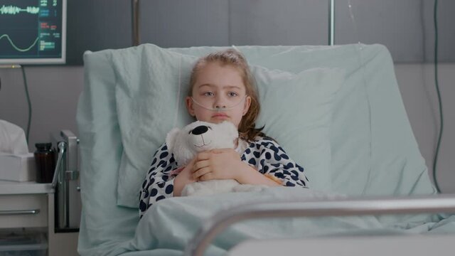 Portrait of lonley sick little child resting in bed holding teedy bear in hands looking at camera during recovery examination in hospital ward. Hospitalized kid waiting for healthcare treatment
