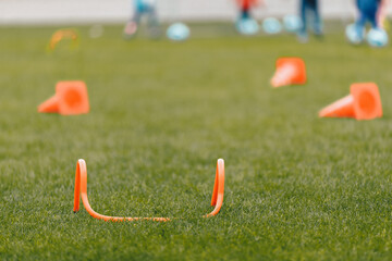 Football Training Equipment on Grass Pitch. Orange Practice Hurdle and Training Cones. Players Running Balls in Blurred Background