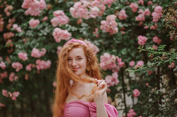 Happy smiling redhead freckled woman plays with her long natural curly hair. Model posing in blooming roses garden, wearing pink headband, blouse. Summer lifestyle, fashion, beauty concept. Copy space