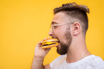 Young handsome bearded man enjoying eating a burger on a yellow background.