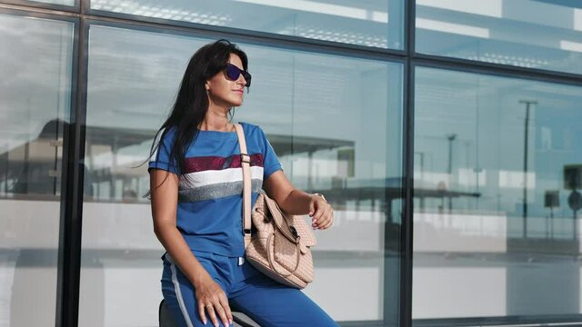 Travel woman sitting on suitcase looking for watch waiting departure at airport terminal exterior