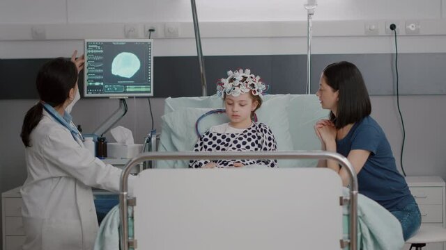 Pediatrician woman doctor monitoring sickness evolution while analyzing brain tomography expertise during neurology consultation. Sick child wearing eeg brain sensors headset in hospital ward