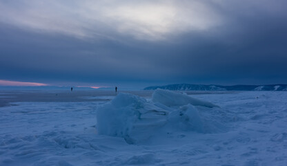 Snowdrifts and ice hummocks lie on the endless frozen lake. In the distance, against the backdrop of a cloudy sunset sky, tiny silhouettes of people walking. Mountains on the horizon. Baikal