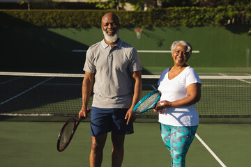 Portrait of smiling senior african american couple with tennis rackets on tennis court