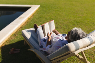 Senior african american woman reading book in deckchair by swimming pool in sunny garden