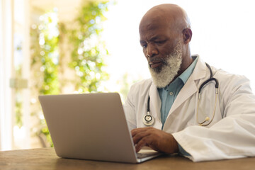 Fototapeta premium Portrait of senior african american male doctor in lab coat using laptop