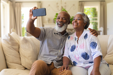 Senior african american couple sitting on sofa using smartphone taking selfies - Powered by Adobe