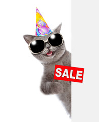 Laughing cat wearing sunglasses and  birthday cap looks from behind empty white banner and holds sales symbol. isolated on white background