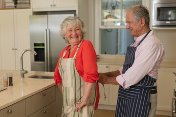 Senior caucasian couple tying apron and smiling in kitchen