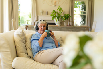 Senior caucasian woman sitting on sofa wearing headphones and holding mug