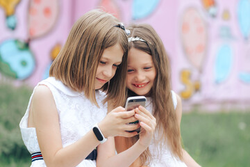Two funny girls in white dresses are watching something on the phone and smiling
