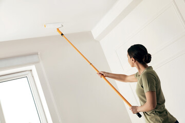 Young woman painting ceiling with white dye indoors, space for text