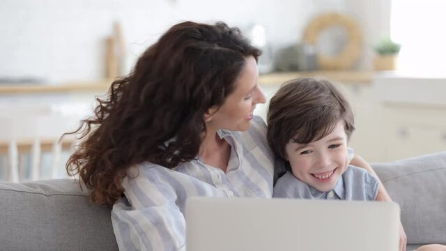 Caring mother spend time with preschool son: mum and kid sit cuddling on sofa use laptop watch cartoon or funny video, make conference video call to friends at home on weekend or coronavirus lockdown