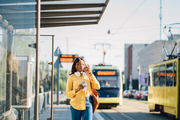 Attractive african american female traveler or student waiting for public transport standing on bus stop outdoor at sunny morning with transport on blurred background.