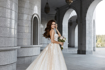 Fototapeta premium A beautiful young bride wearing a luxurious lace bridal dress posing outdoors. Outdoor romantic portrait of an attractive brunette girl with bright makeup and wedding hairstyle
