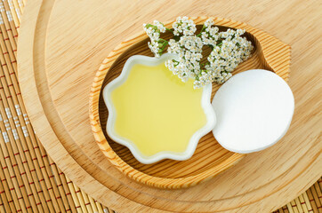 Small white bowl with cosmetic (cleansing) aroma oil, cotton pad and white yarrow flowers. Natural skin care, homemade spa and beauty treatment recipe. Top view, copy space.