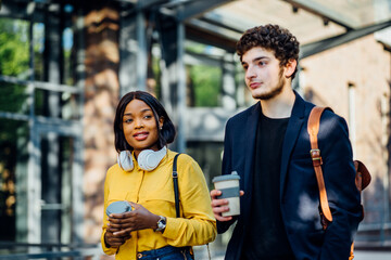Multiethnic couple talking to each other and drinking coffee while walking in the city. Millennial african female and french male students together outdoor at sunny summer day.