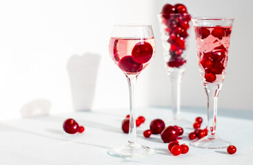 set of glasses of pink gin or vodka infused with cranberry among frozen berries, a long-stemmed crystal glasses with cherry liqueur or any red alcoholic cocktail on light blue background, copy space