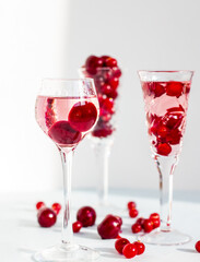 Fototapeta na wymiar set of glasses of pink gin or vodka infused with cranberry among frozen berries, a long-stemmed crystal glasses with cherry liqueur or any red alcoholic cocktail on light blue background, minimalism