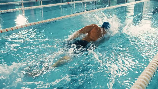 Male Swimmer Swimming in Olympic Pool. Professional Athlete Performing at Championship, using Front Crawl, Freestyle Technique. Determination to Win. High Angle Cinematic Tracking Slow Motion