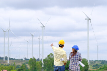 An engineer stands holding a blueprint and looks at a wind turbine project to generate electricity and check wind direction.